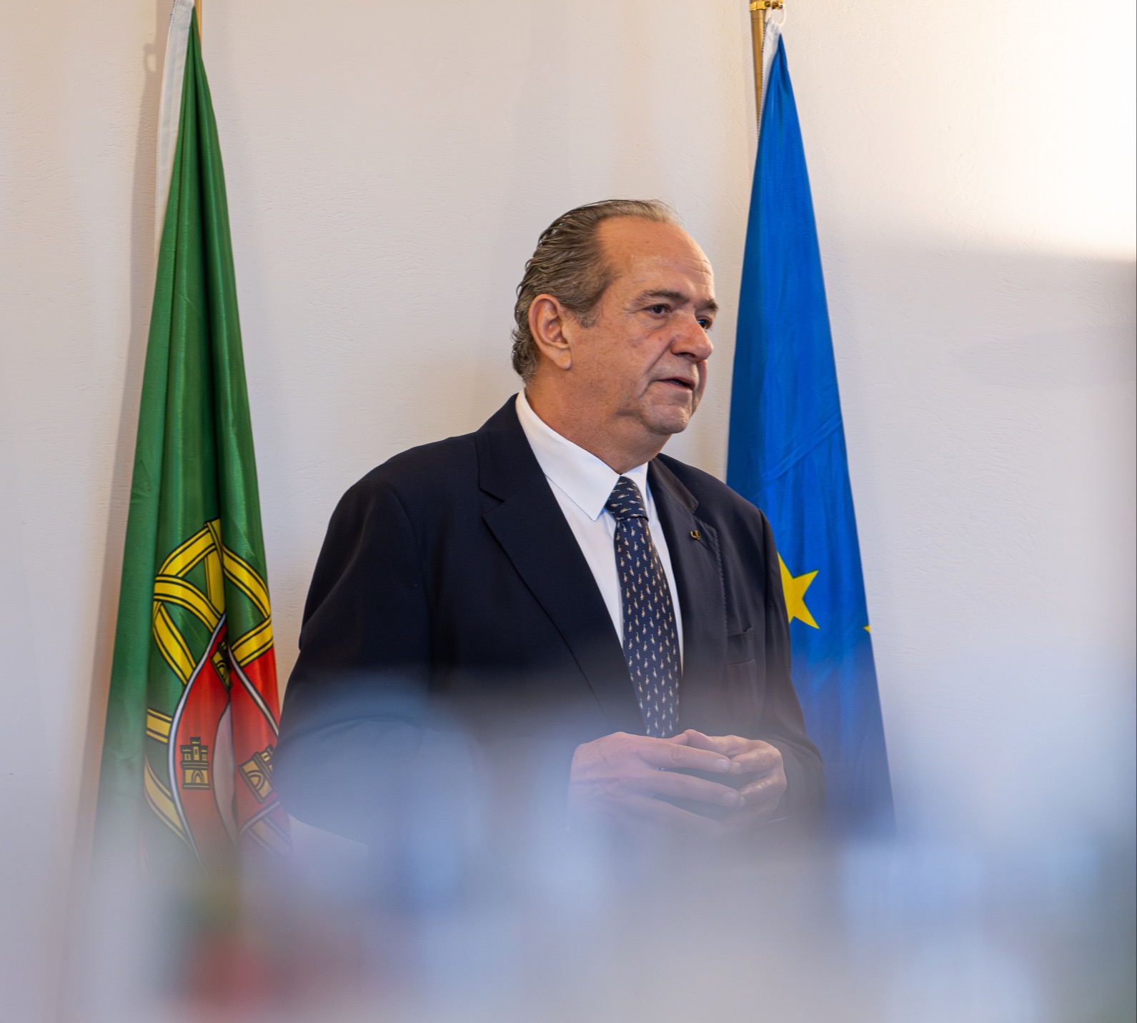 Pessoa em fato e gravata a discursar em frente às bandeiras de Portugal e da União Europeia.