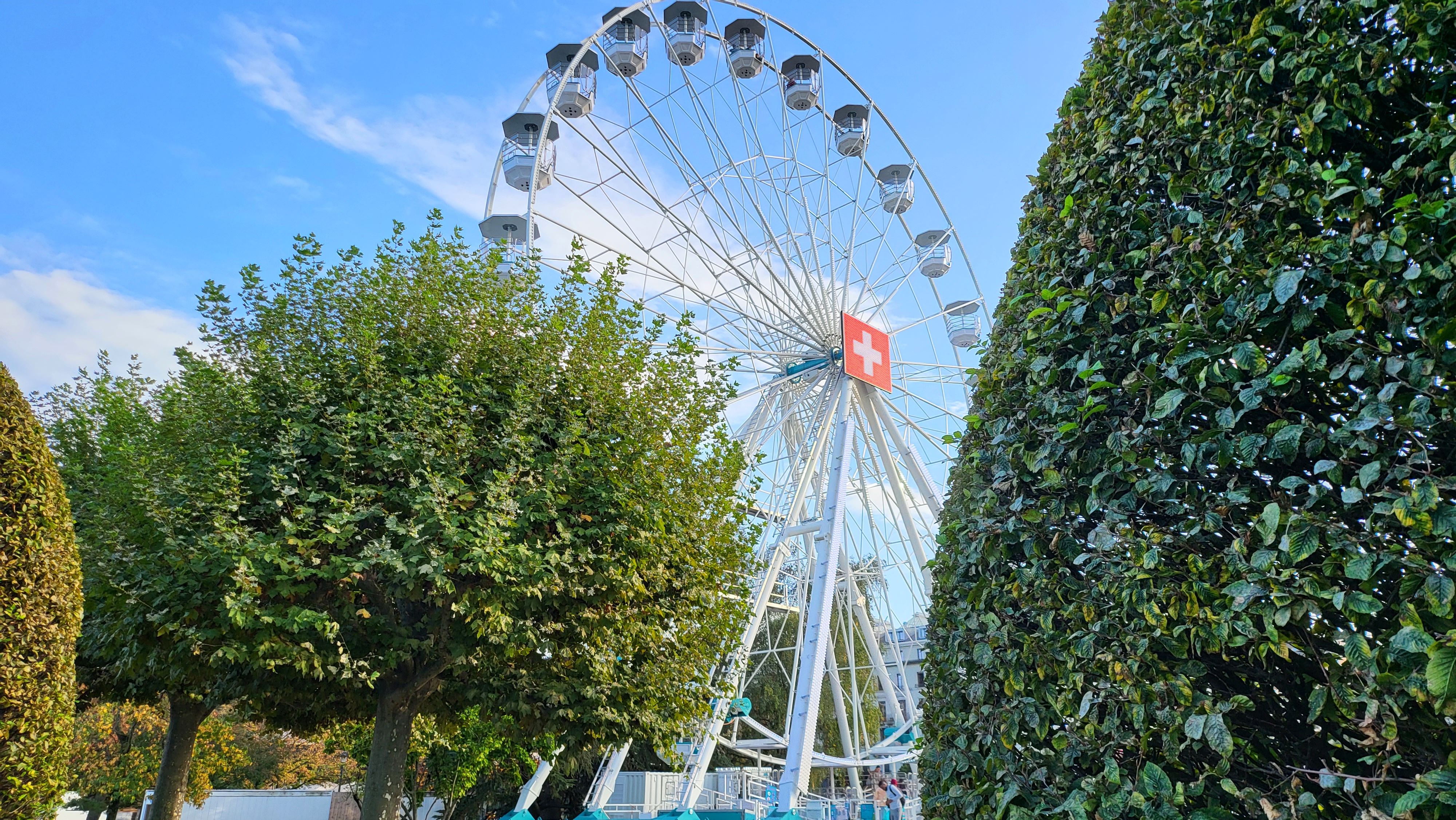 Roda-gigante visível por entre árvores bem cuidadas num parque, sugerindo um ambiente público na Suíça.