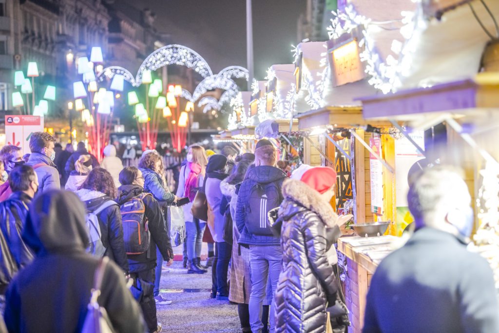 Mercado de Natal em Bruxelas à noite, com barracas iluminadas, luzes festivas e muitas pessoas a passear, algumas com guarda-chuva, criando um ambiente animado e acolhedor.