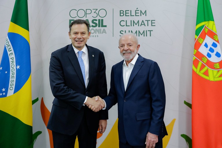 O presidente do Brasil e o primeiro-ministro de Portugal apertam as mãos, sorridentes, em frente a painéis com os logótipos da Cúpula de Líderes de Belém e as bandeiras do Brasil e de Portugal ao fundo.