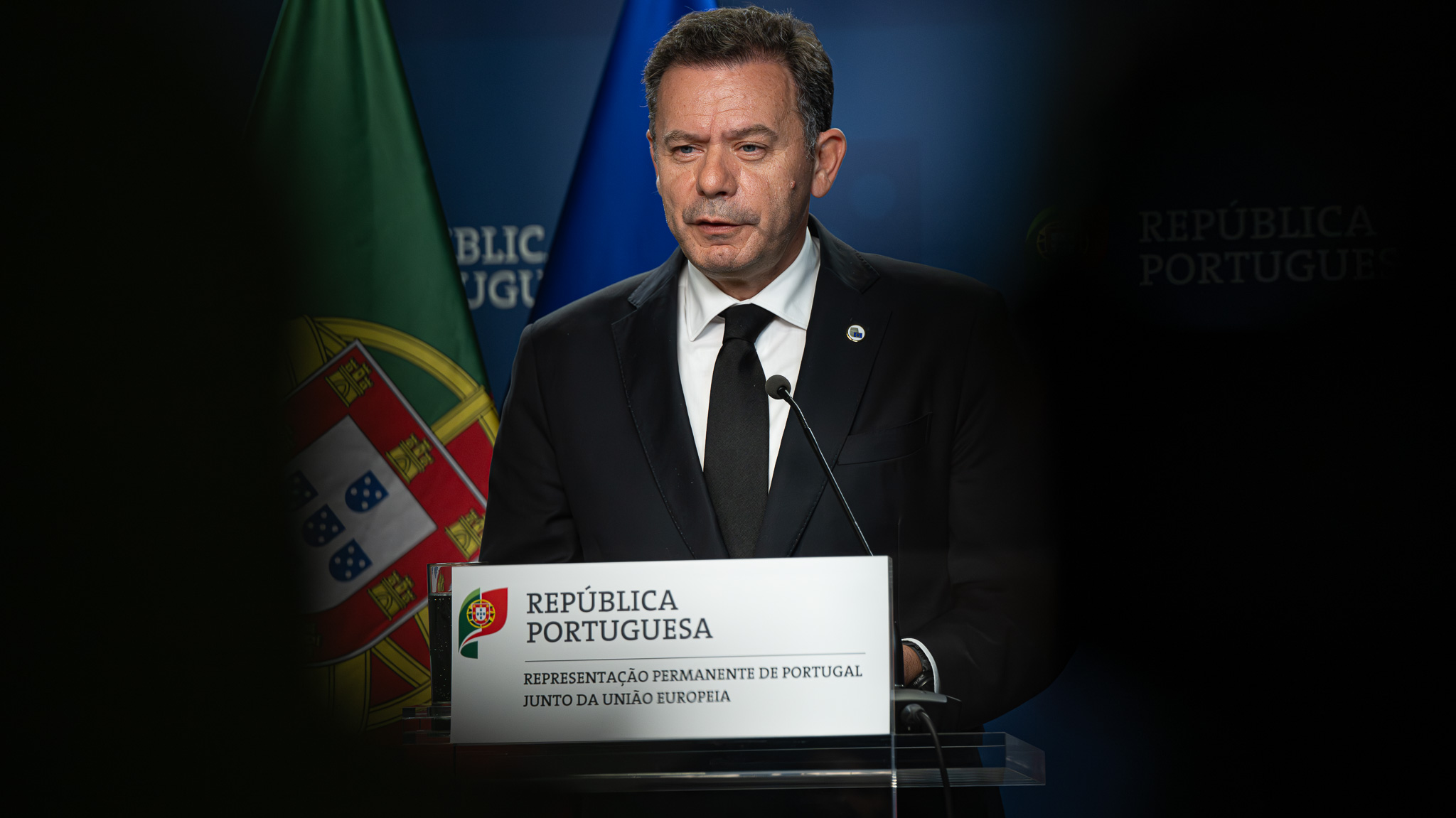 O primeiro-ministro português, Luís Montenegro, fala numa conferência de imprensa oficial, com a bandeira de Portugal ao fundo e um púlpito com o logótipo da República Portuguesa.