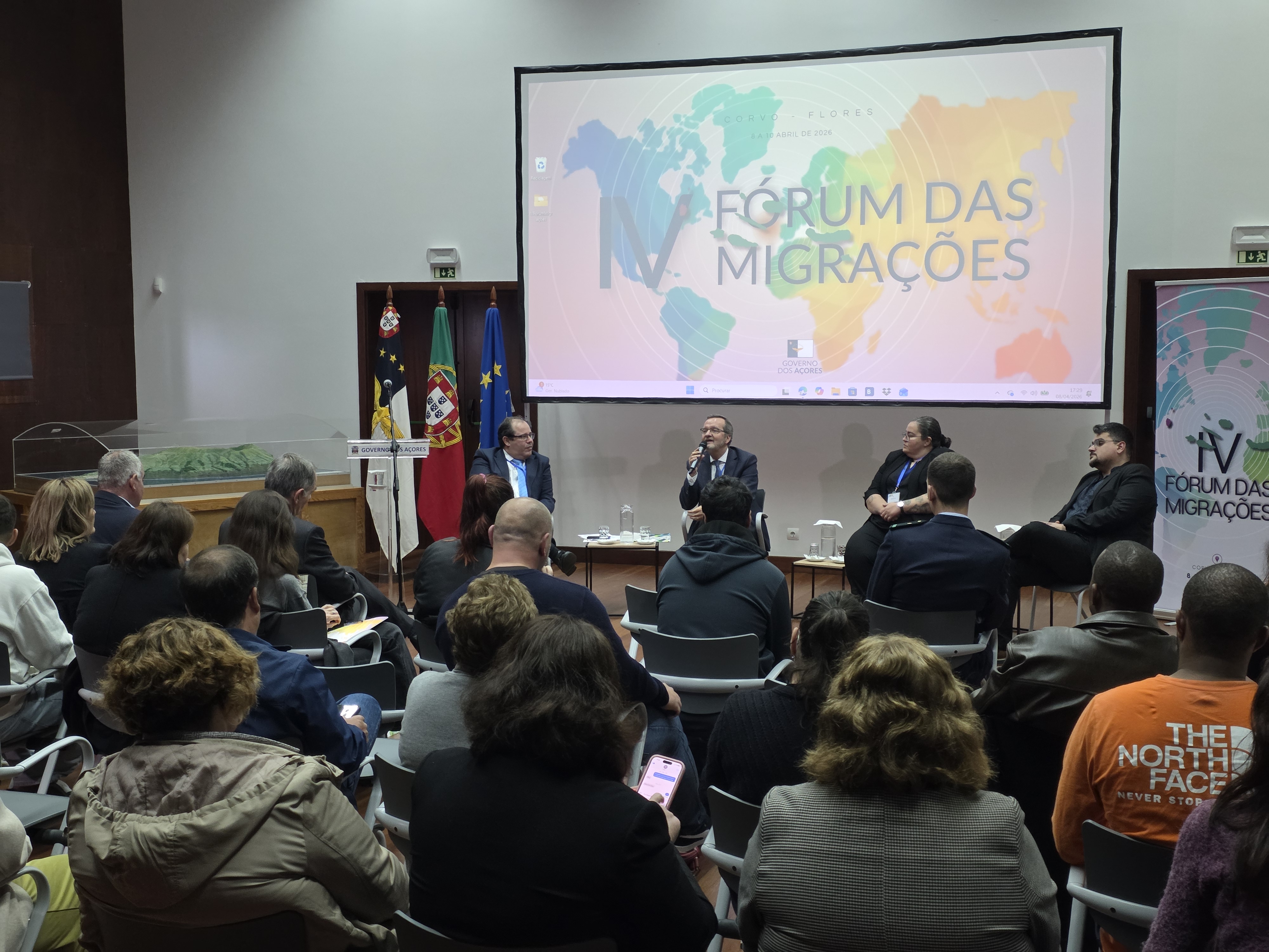 Auditório com público sentado a assistir a uma mesa-redonda de quatro oradores, com um ecrã ao fundo que exibe o título 'Fórum das Migrações' e um mapa-mundo colorido, num evento dedicado ao debate sobre migrações nos Açores.