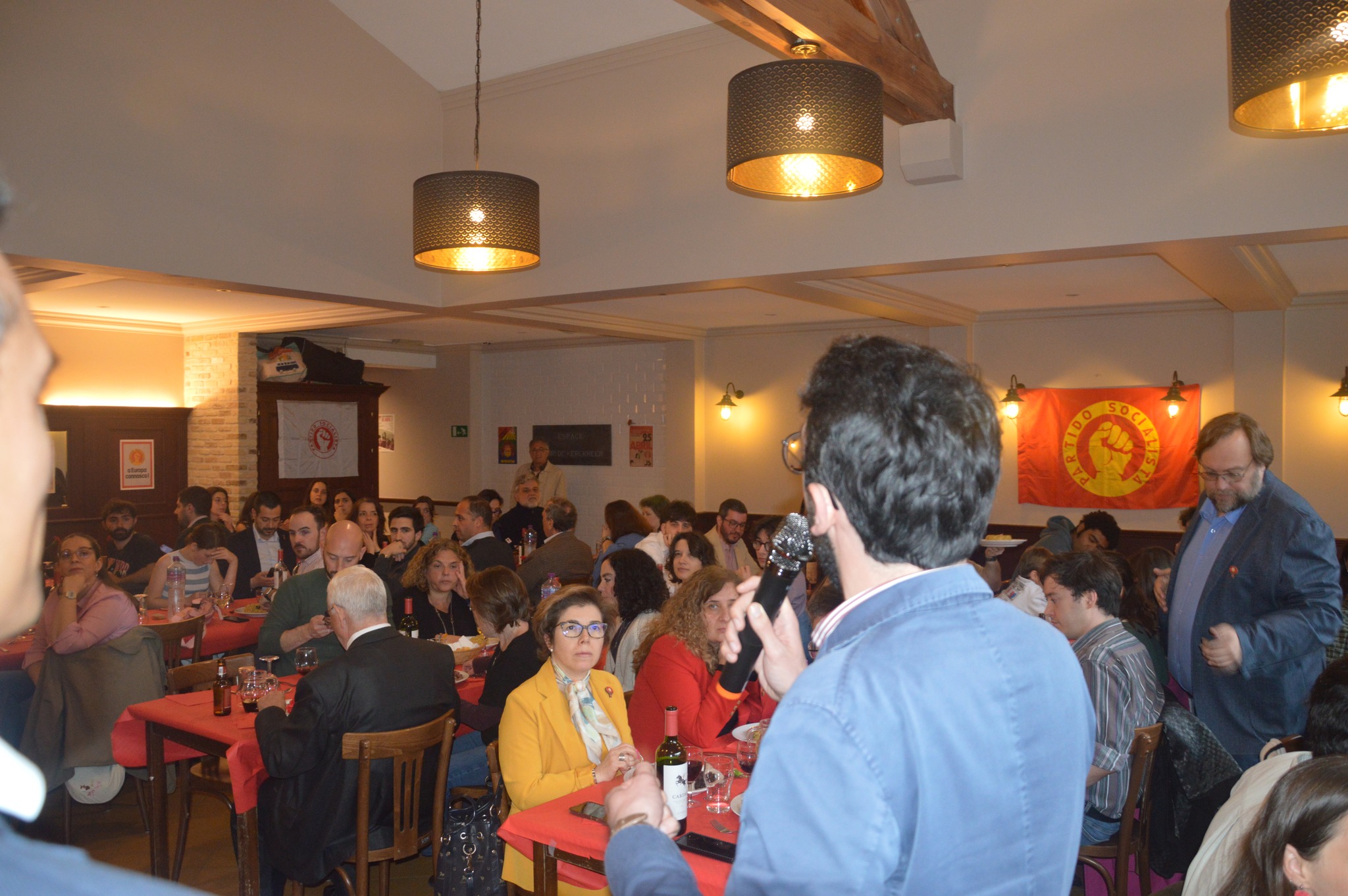 Jantar solidário com várias pessoas sentadas em mesas, enquanto uma pessoa discursa ao microfone; decoração com símbolos do Partido Socialista visível na parede, refletindo um ambiente de celebração e solidariedade.