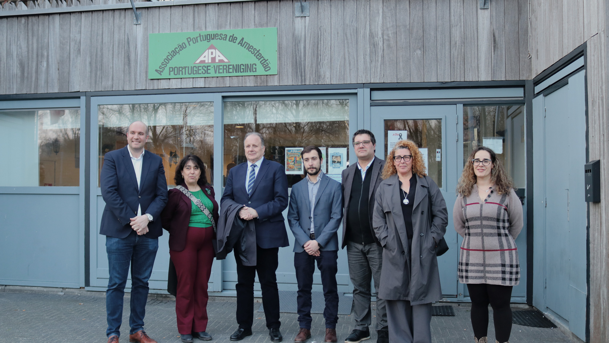 Sete pessoas posam em frente à entrada do Grupo Desportivo Casa Portuguesa de Haia, ilustrando a visita de representantes da comunidade portuguesa nos Países Baixos.