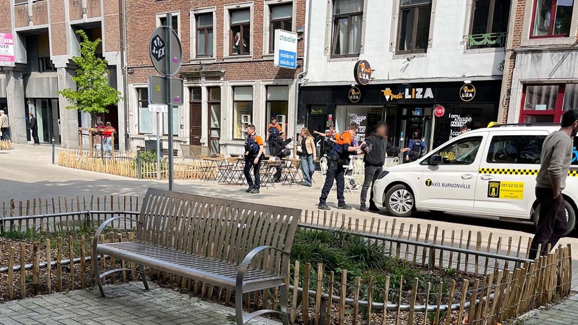 Polícias e uma viatura policial numa rua do centro de Namur, em frente a edifícios comerciais, com uma área isolada e pessoas a observar, ilustrando a intervenção após um tiroteio no bairro universitário.