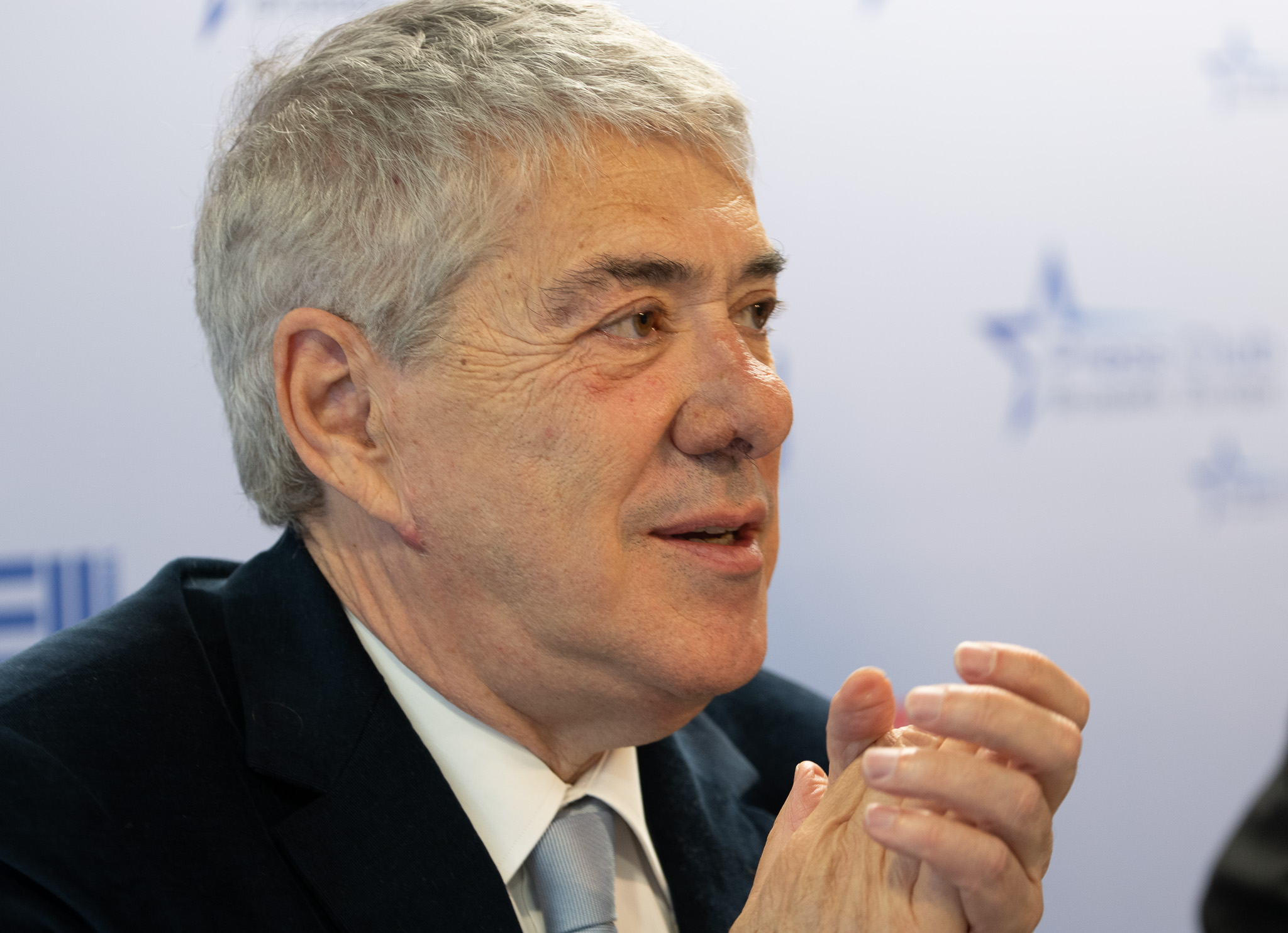 Pessoa de cabelo grisalho, em fato e gravata, sentada num ambiente formal, aparentemente a falar ou ouvir durante uma conferência de imprensa.