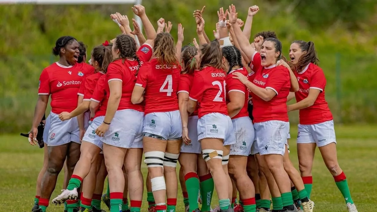 Grupo de jogadoras da Seleção Nacional Feminina de Rugby de Portugal, com equipamento vermelho e branco, a celebrar juntas em círculo no campo, com os braços erguidos, mostrando união e entusiasmo antes de um jogo importante.