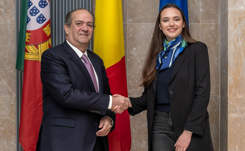 O Embaixador de Portugal na Bélgica e a Ministra federal belga das Classes Médias, Independentes e PME apertam as mãos num ambiente formal, com as bandeiras de Portugal e Bélgica ao fundo, simbolizando cooperação bilateral.