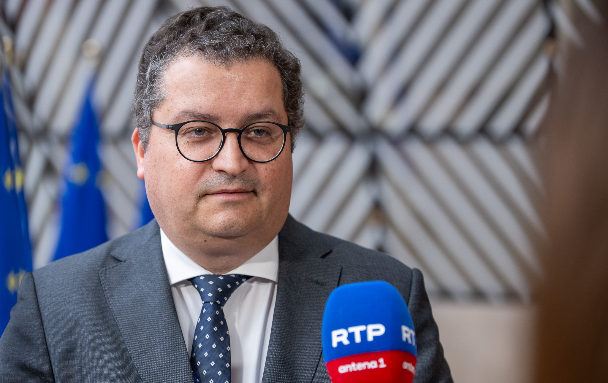 O ministro das Finanças português, de fato e gravata, fala à imprensa em Bruxelas diante de bandeiras da União Europeia, com um microfone da RTP à sua frente.