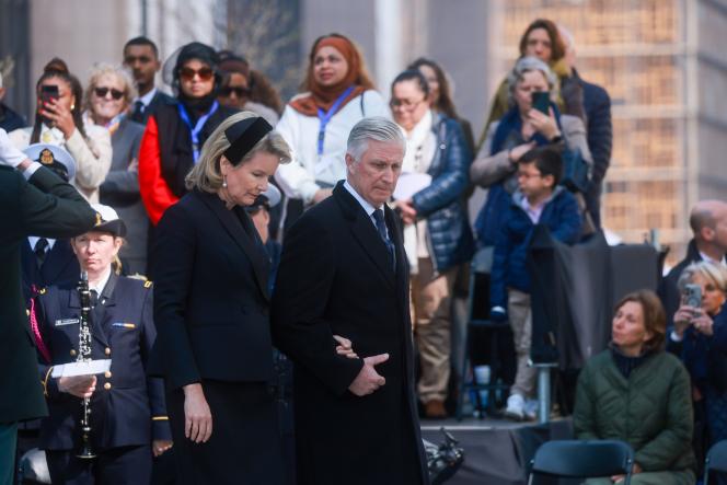 O Rei Philippe caminha de braço dado com uma mulher, ambos com expressão solene, durante uma cerimónia pública de homenagem. Atrás deles, várias pessoas assistem em silêncio, refletindo o ambiente de respeito e memória pelas vítimas dos atentados de 22 de março de 2016 em Bruxelas.