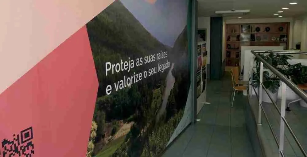 Corredor interior com uma parede decorada com a frase 'Proteja as suas raízes e valorize o seu legado', acompanhada de uma imagem de paisagem natural e um QR code no canto inferior esquerdo.