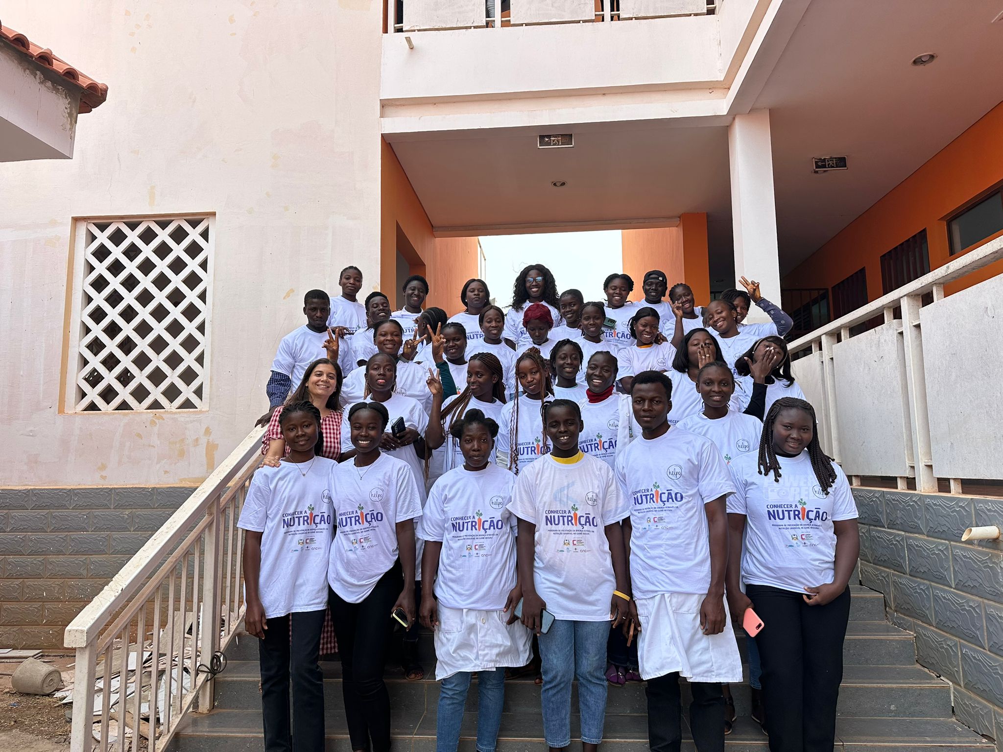 Grupo de pessoas, maioritariamente jovens adultos, vestindo t-shirts brancas alusivas ao projeto de licenciatura em Dietética e Nutrição, posam numa escadaria em frente a um edifício escolar, simbolizando a colaboração académica entre Portugal e Guiné-Bissau.