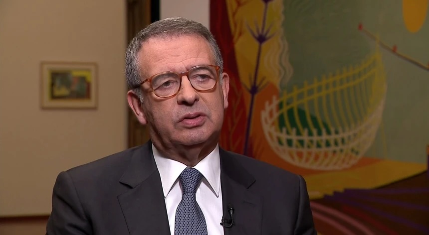 António José Seguro, de fato e gravata, durante uma entrevista formal, com uma pintura colorida ao fundo, ilustrando o contexto político e institucional da conversa sobre as comunidades portuguesas no estrangeiro.