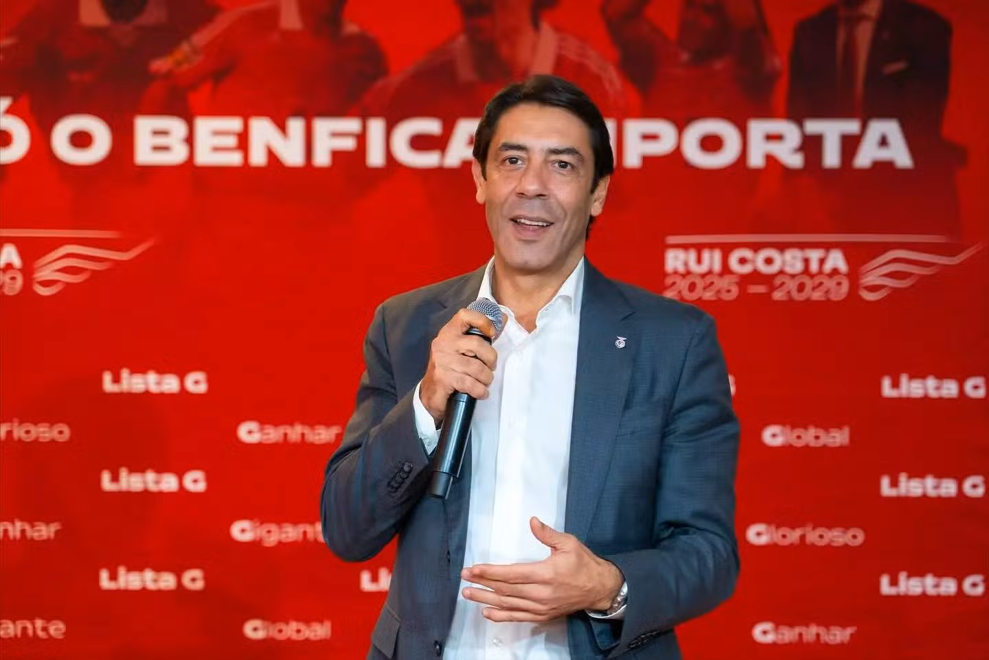 Rui Costa, de fato escuro e camisa branca, discursa com um microfone à frente de um fundo vermelho com referências ao Benfica e ao processo eleitoral do clube.