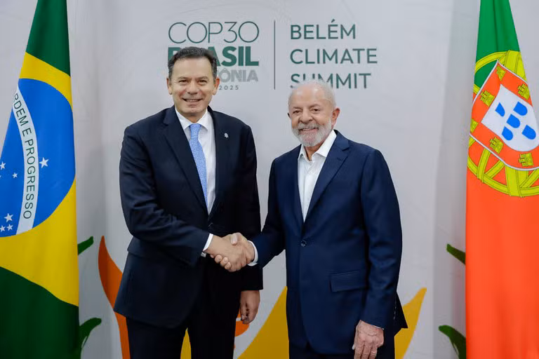 O presidente do Brasil e o primeiro-ministro de Portugal apertam as mãos, sorridentes, em frente a painéis com os logótipos da Cúpula de Líderes de Belém e as bandeiras do Brasil e de Portugal ao fundo.