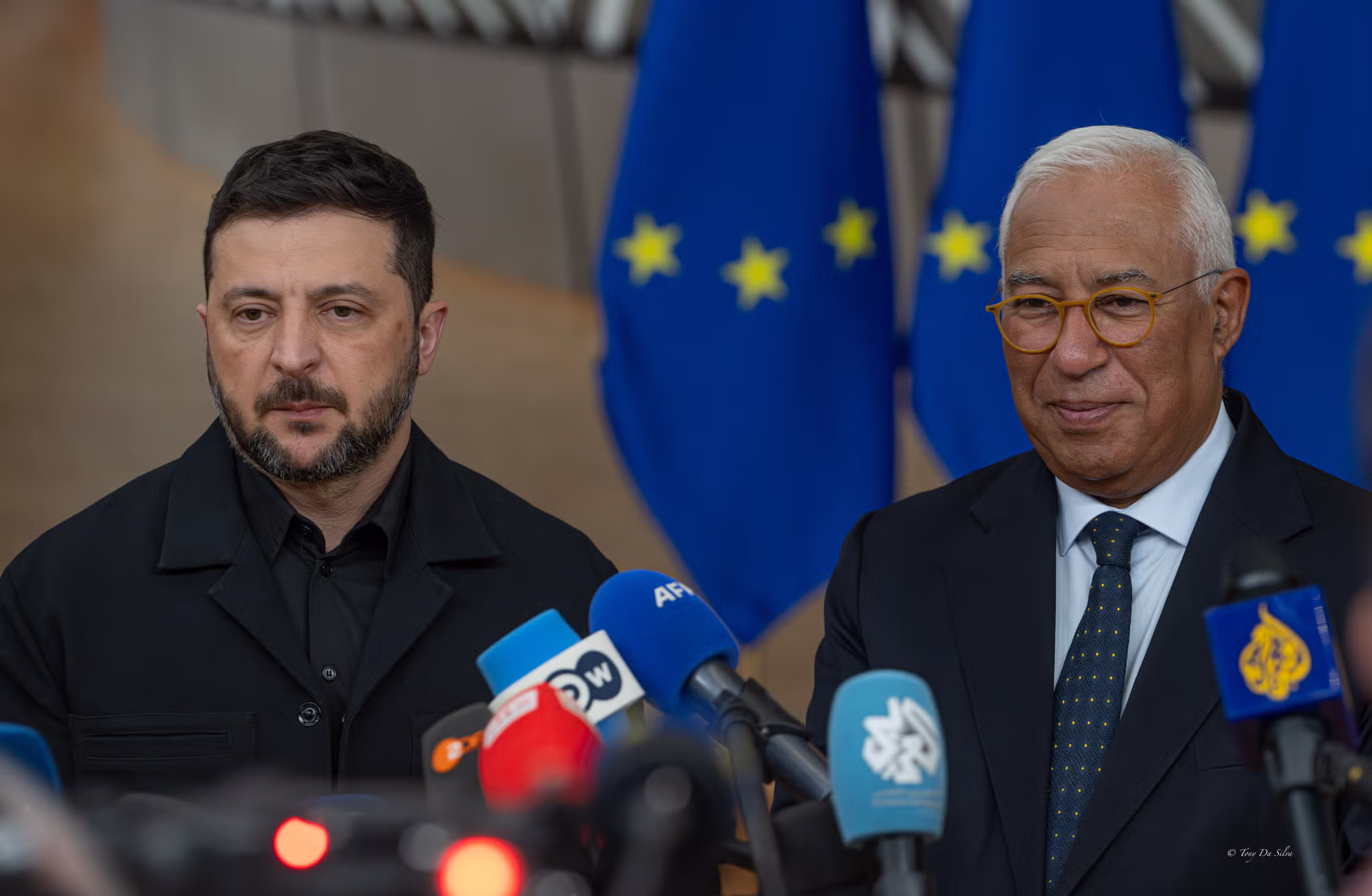 Dois líderes políticos falam à imprensa em frente a várias bandeiras da União Europeia, rodeados de microfones, durante uma cimeira europeia em Bruxelas sobre o apoio à Ucrânia.