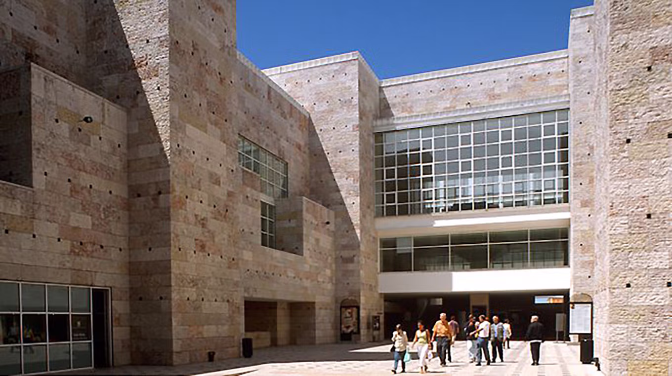 Fachada moderna do Centro Cultural de Belém, com paredes de pedra clara, grandes janelas e um grupo de pessoas junto à entrada, sob céu azul.