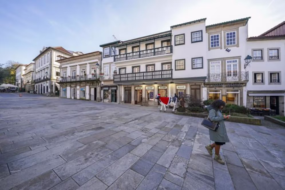 Praça urbana com edifícios comerciais de fachadas tradicionais, lojas no rés-do-chão e uma pessoa a caminhar em primeiro plano.
