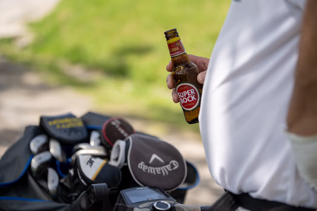 Pessoa com camisa branca segura uma garrafa de cerveja ao lado de um saco de tacos de golfe, sugerindo um momento informal durante um evento de golfe.
