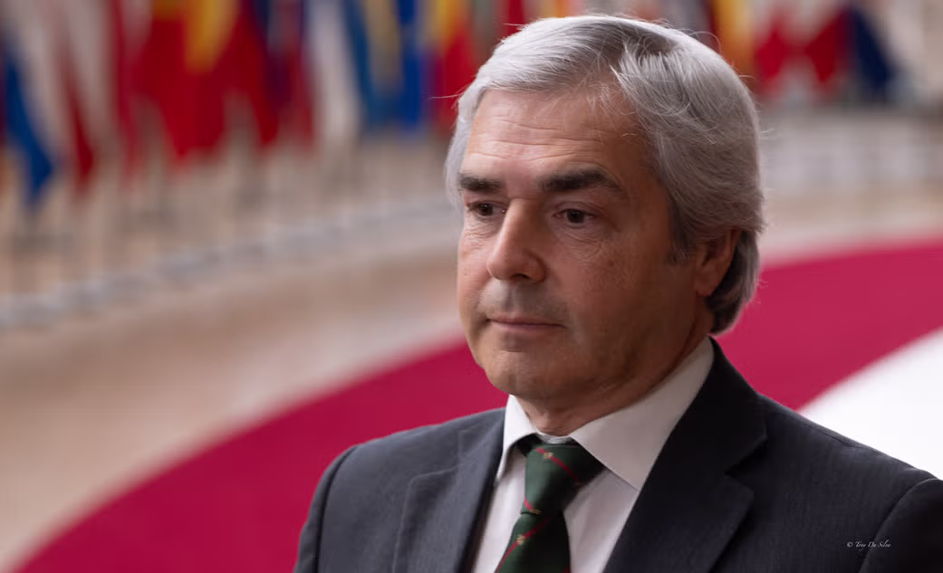 Homem de fato escuro e gravata, em ambiente institucional com várias bandeiras ao fundo, ilustrando uma reunião política europeia.