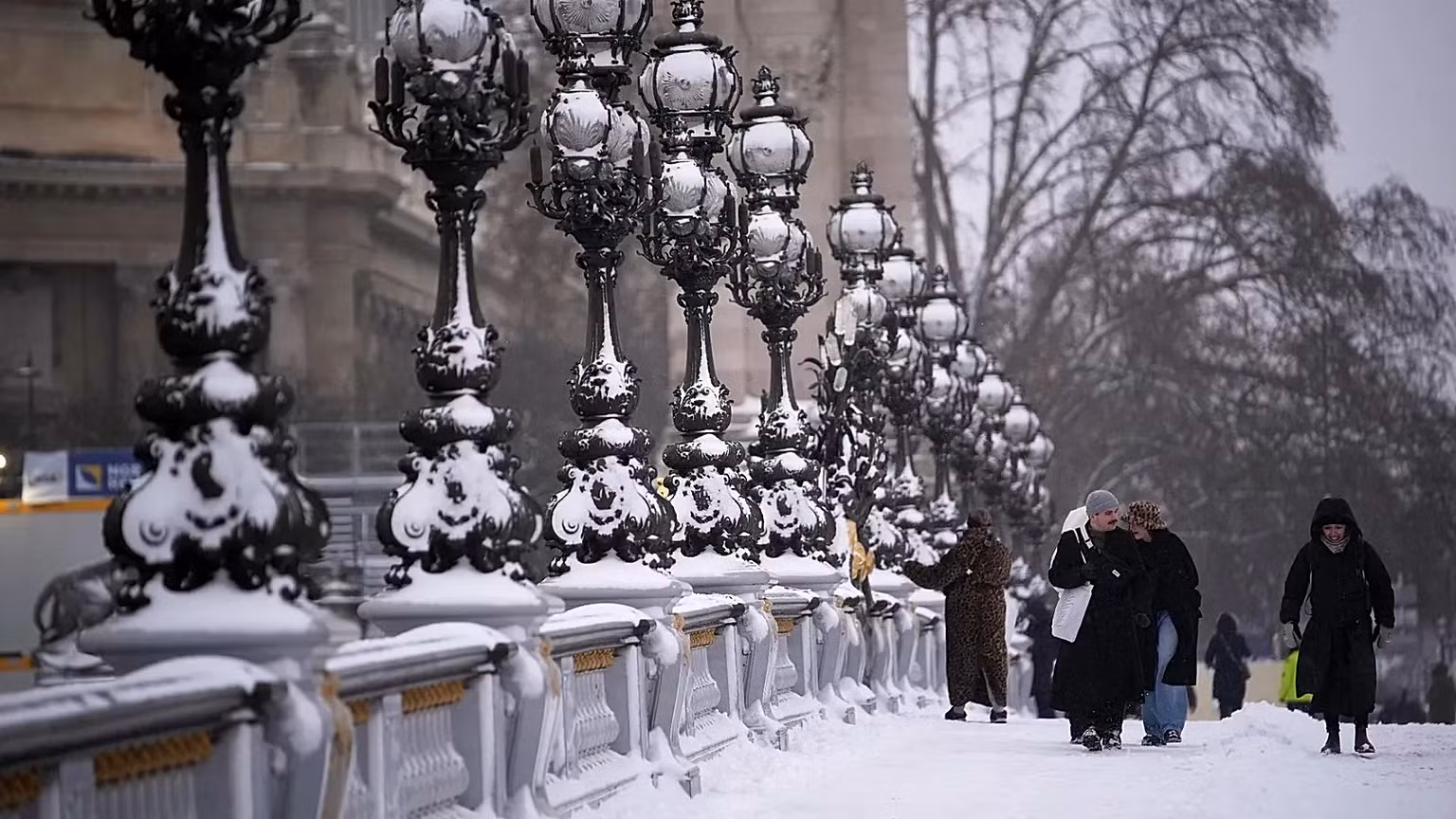 Passeio urbano coberto de neve com candeeiros ornamentados e várias pessoas a caminhar, num ambiente de inverno.