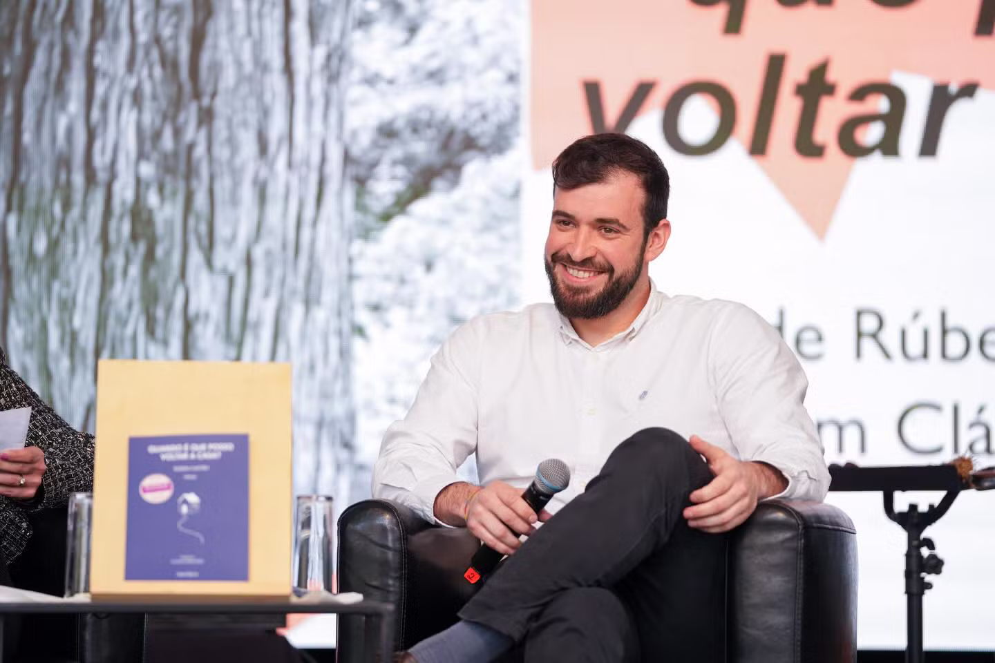 Rúben Castro sentado numa cadeira, sorridente, com um microfone na mão, durante a apresentação do seu livro de poesia 'Quando é que posso voltar a casa?', com o livro exposto numa mesa à sua frente.