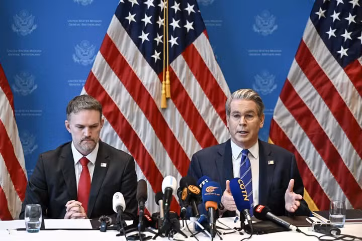 Conferência de imprensa com dois homens em fato e gravata sentados à mesa, rodeados por microfones, com bandeiras dos Estados Unidos ao fundo, representando uma comunicação oficial do governo norte-americano.