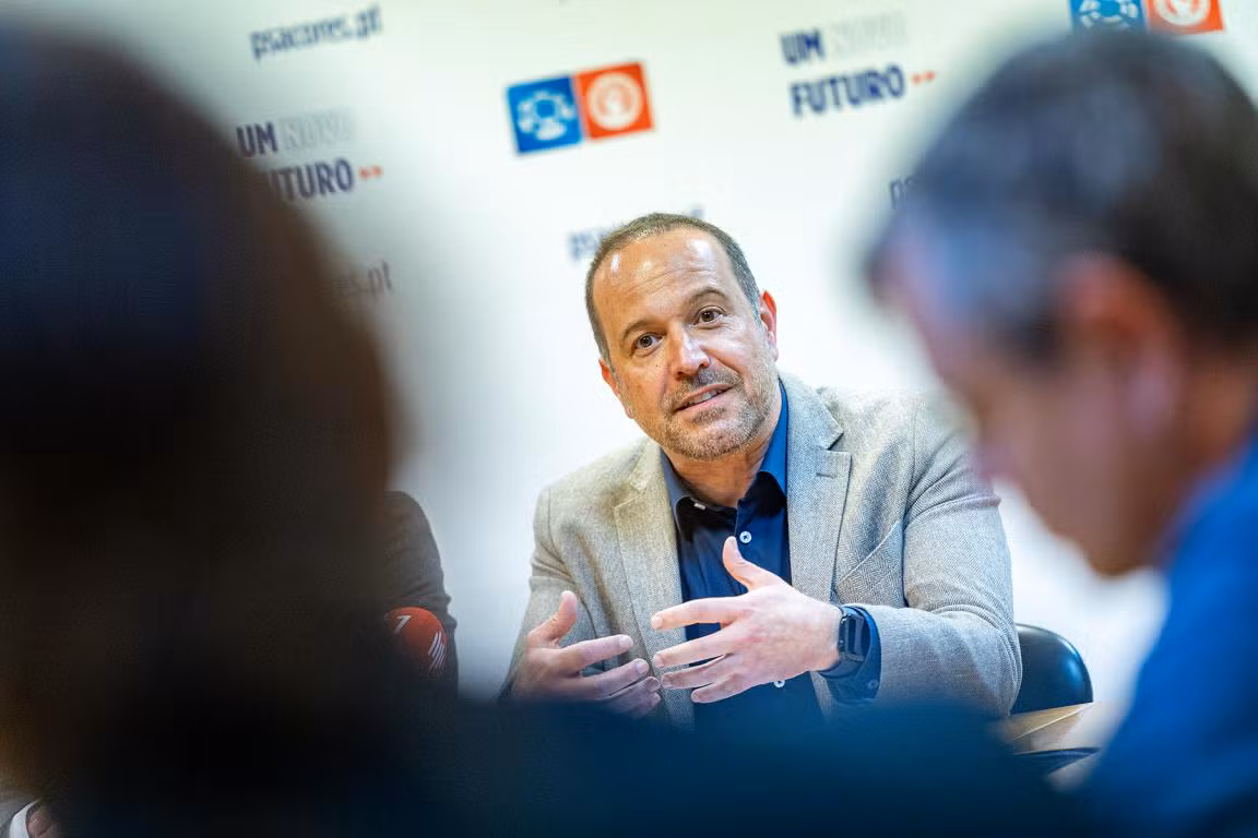 Francisco César, líder do PS/Açores, fala gesticulando numa reunião formal, com fundo decorado com logótipos e frases do partido, acompanhado por outras pessoas desfocadas em primeiro plano.