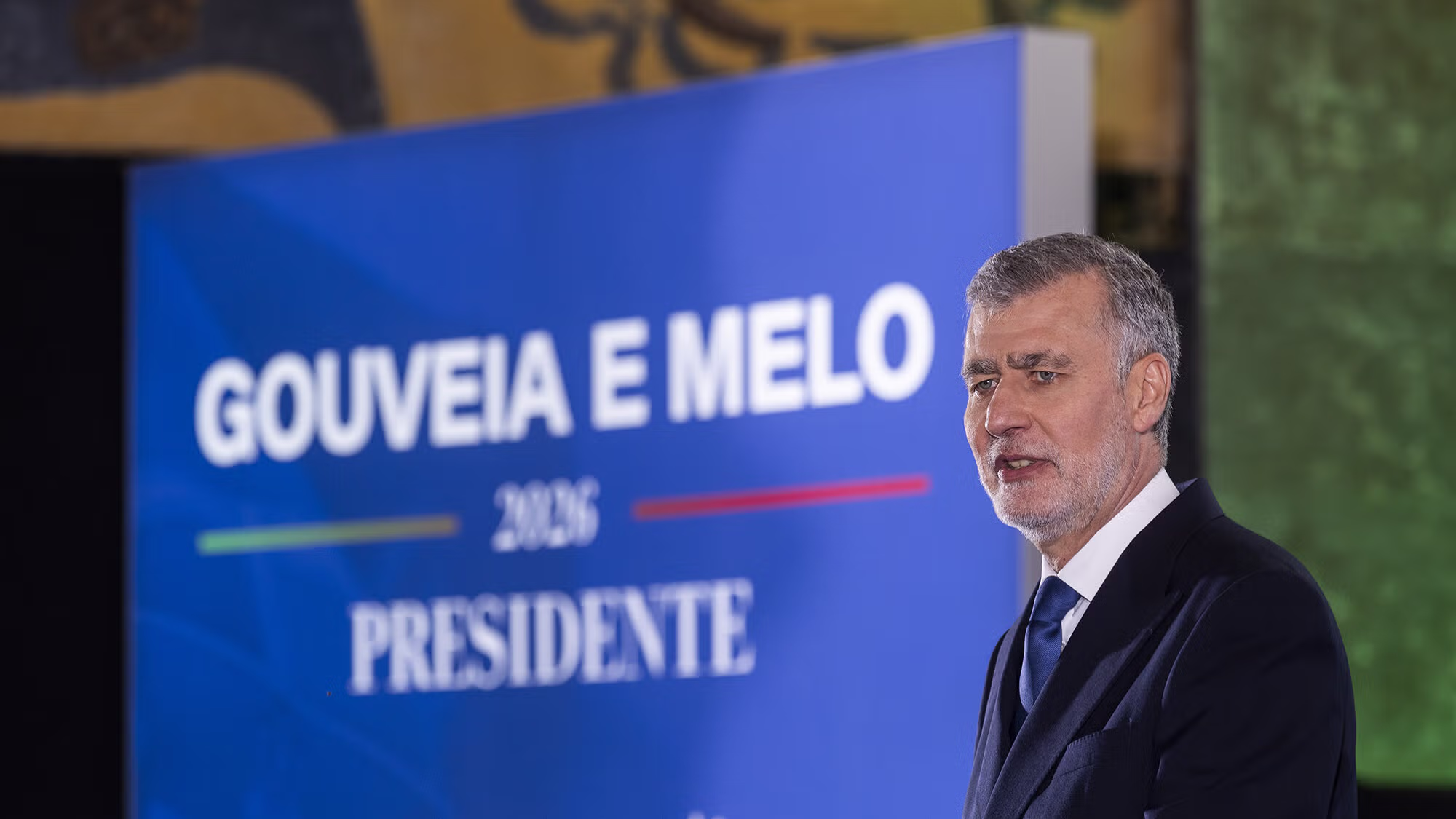 O almirante Gouveia e Melo, de fato escuro, está em frente a um painel azul com o seu nome e a palavra 'Presidente', num cenário de campanha eleitoral para as presidenciais de 2026.