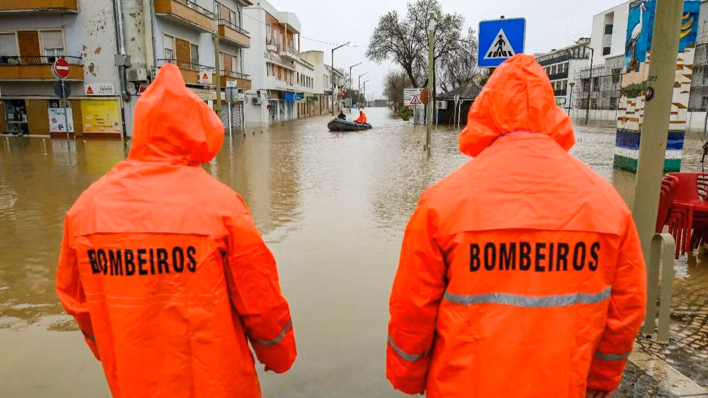 Dois bombeiros com impermeáveis laranja observam uma rua inundada, com uma equipa de resgate num barco ao fundo, ilustrando a resposta de emergência às cheias em Portugal.